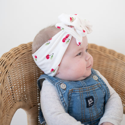 Baby wearing a white headband with cherries print and denim overalls sitting in a wicker chair.