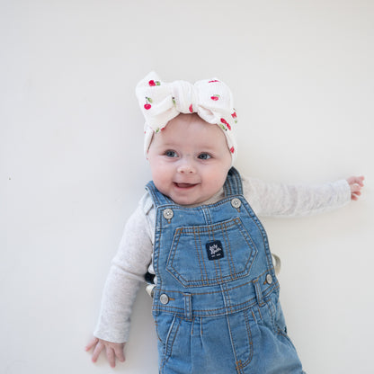 Baby wearing a denim overall outfit with a white and red headband on a plain background