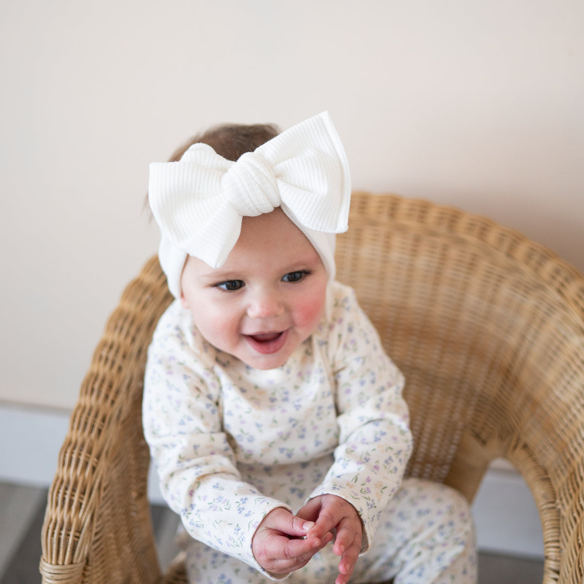 Baby wearing a white headband with a large bow, sitting on a wicker chair.
