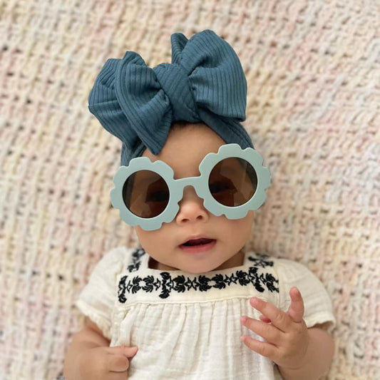 Baby wearing light blue flower sunglasses and a matching blue ribbed oversized bow headband on a textured beige blanket.
