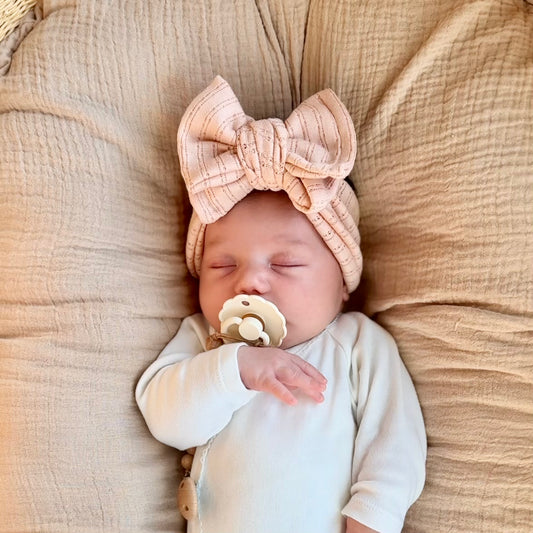 Newborn baby sleeping with a pacifier and pink headband on a beige blanket