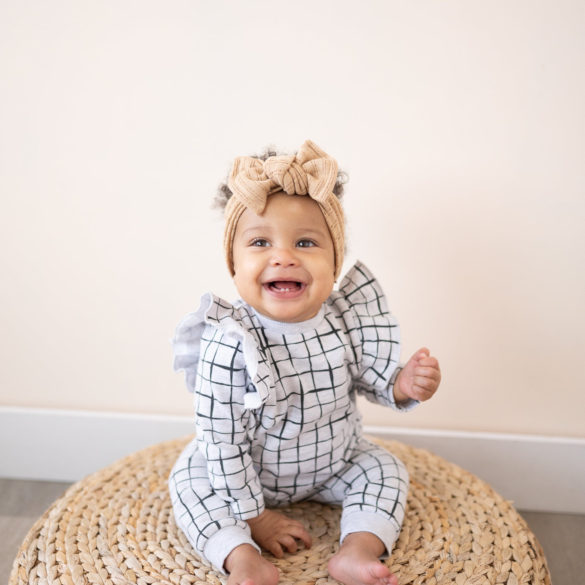 Baby wearing a checkered outfit and a caramel top knot headband with a bow, sitting on a woven cushion.