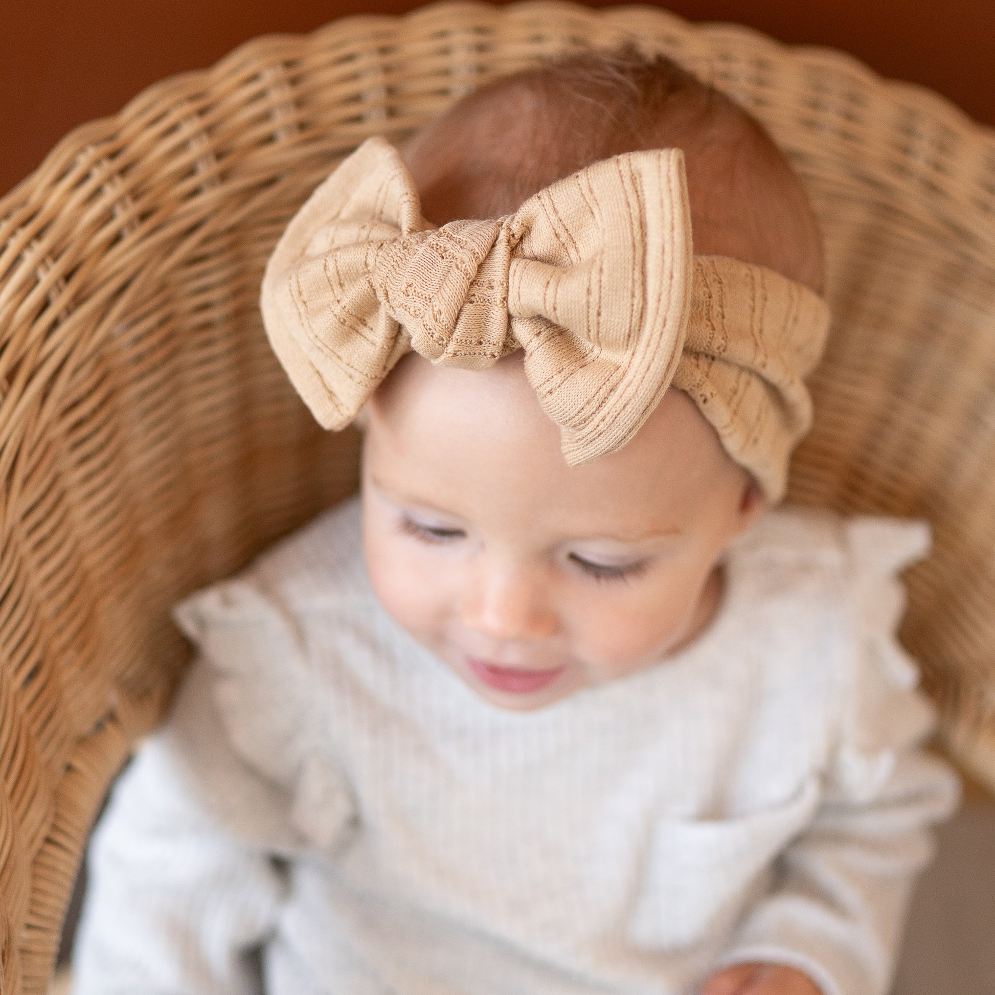 Baby wearing a caramel beige bow headband sitting in a wicker chair.