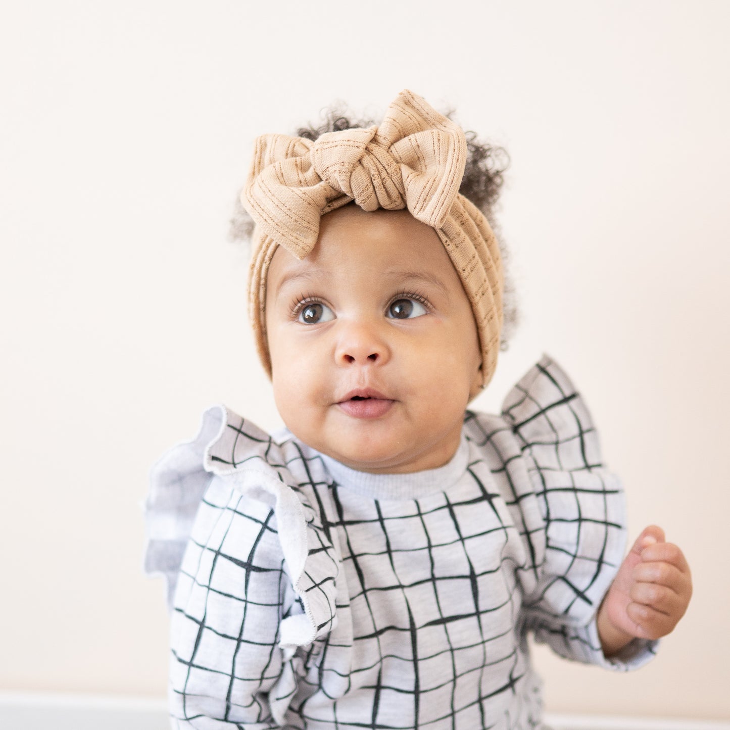 Baby wearing a beige headband with a bow and a checkered outfit on a light background