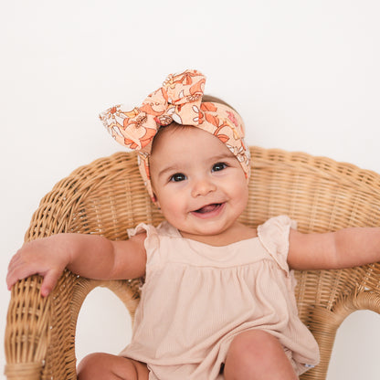 Baby sitting on a wicker chair wearing a floral headband and a light-colored outfit.