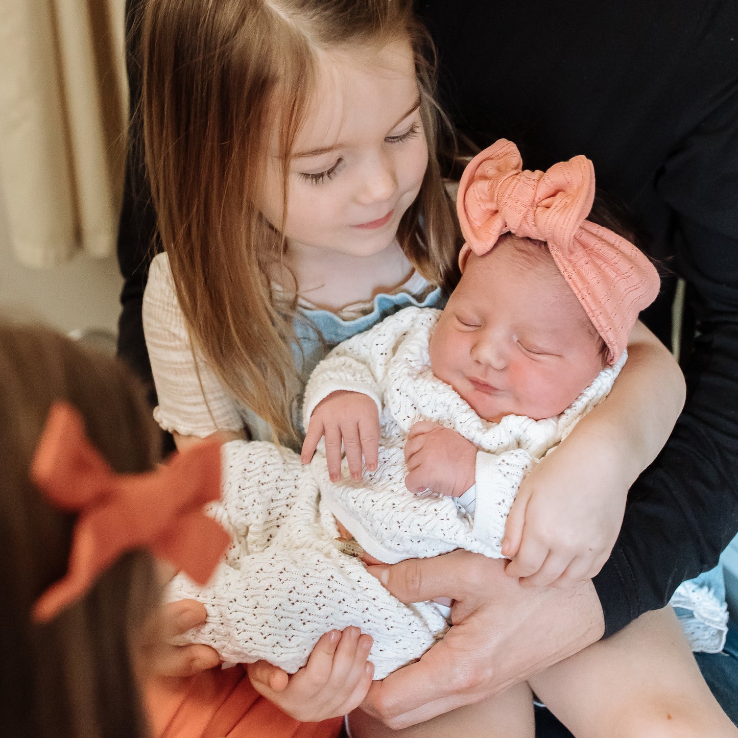 Young girl holding a newborn baby in off white crochet outfit with a coral pointelle top knot bow.