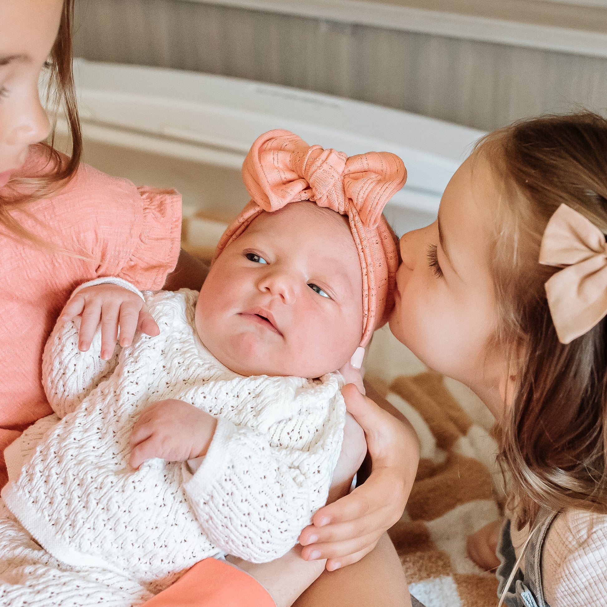 Two older children interacting with a baby wearing a white outfit and coral top knot headband.