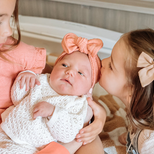 Two older children interacting with a baby wearing a white outfit and coral top knot headband.