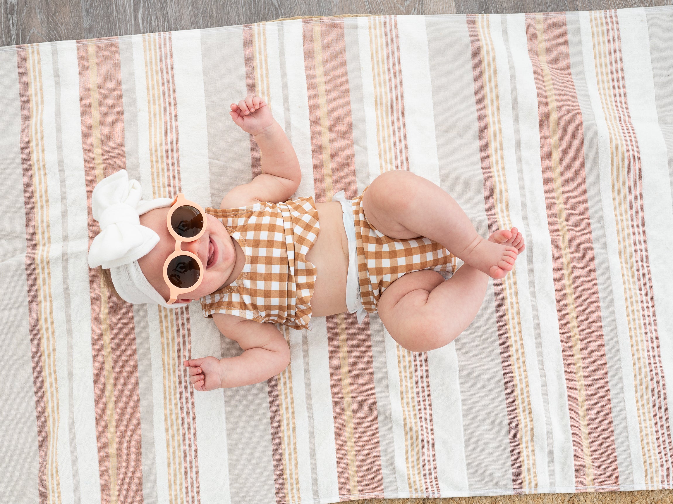 baby wearing brown and white gingham swimsuit with a white oversized bow and pink sunglasses.