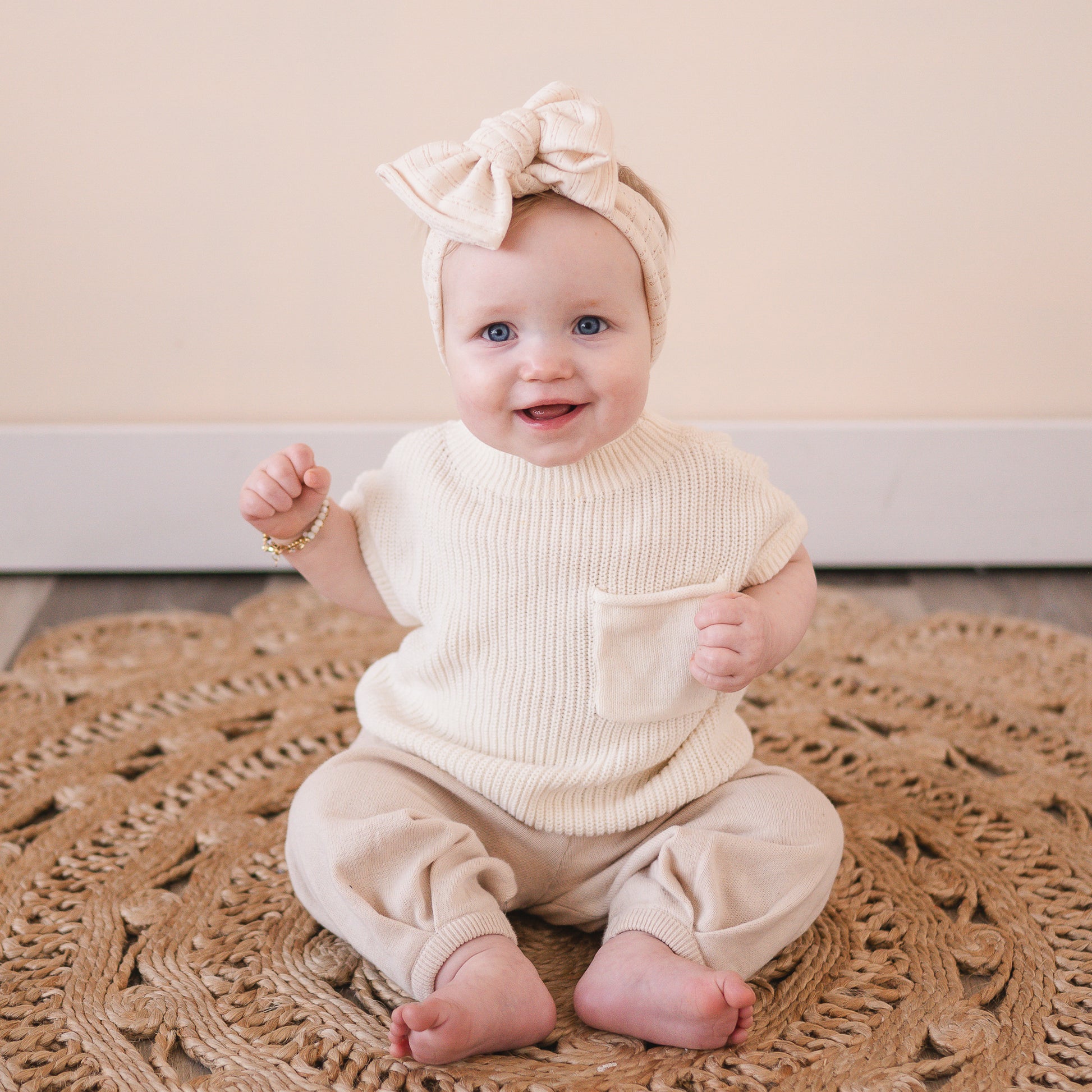 Baby wearing a cream-colored outfit with an oversized pointelle knit bow headband, sitting on a textured rug.