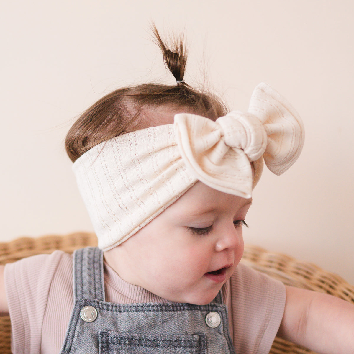 Baby wearing a beige headband with a bow, sitting on a woven surface.