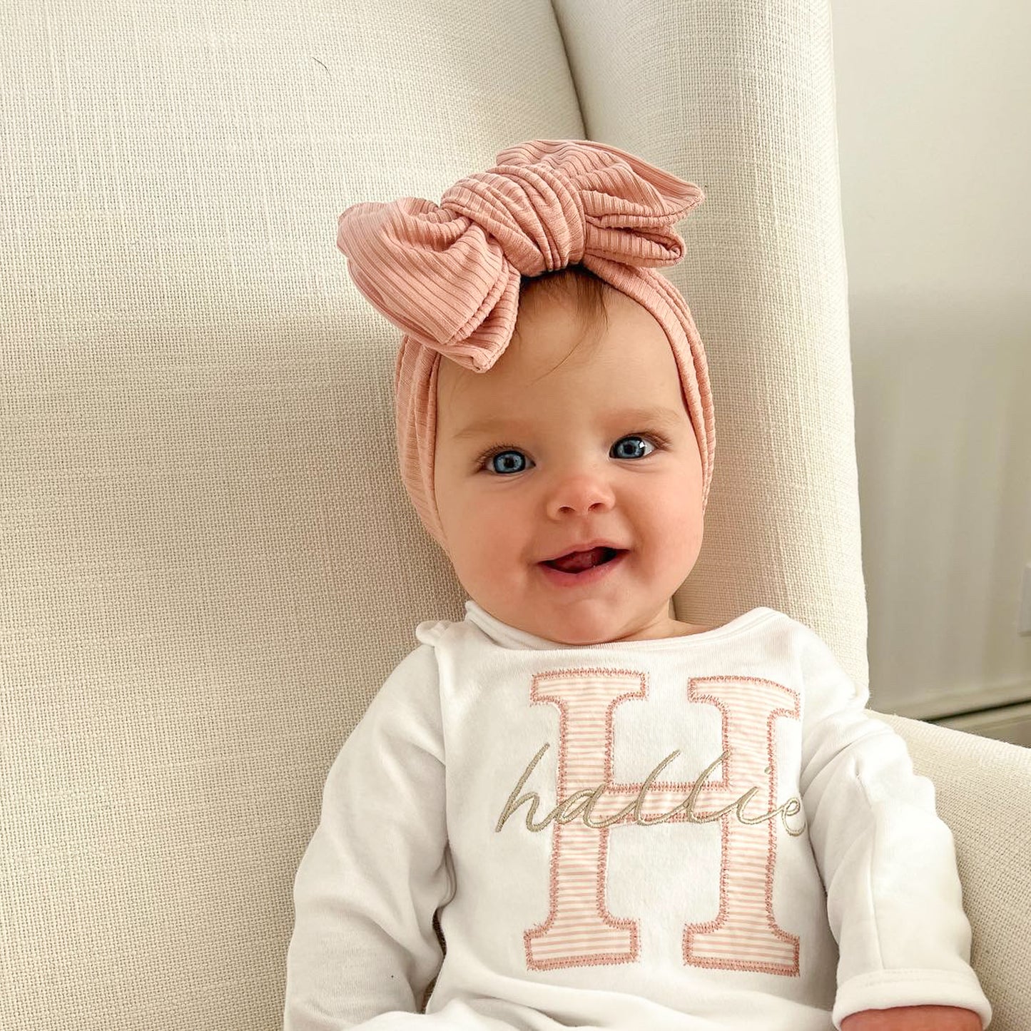Baby wearing a dusty pink headband and a white outfit with 'Hattie' printed on it, sitting on a beige couch.
