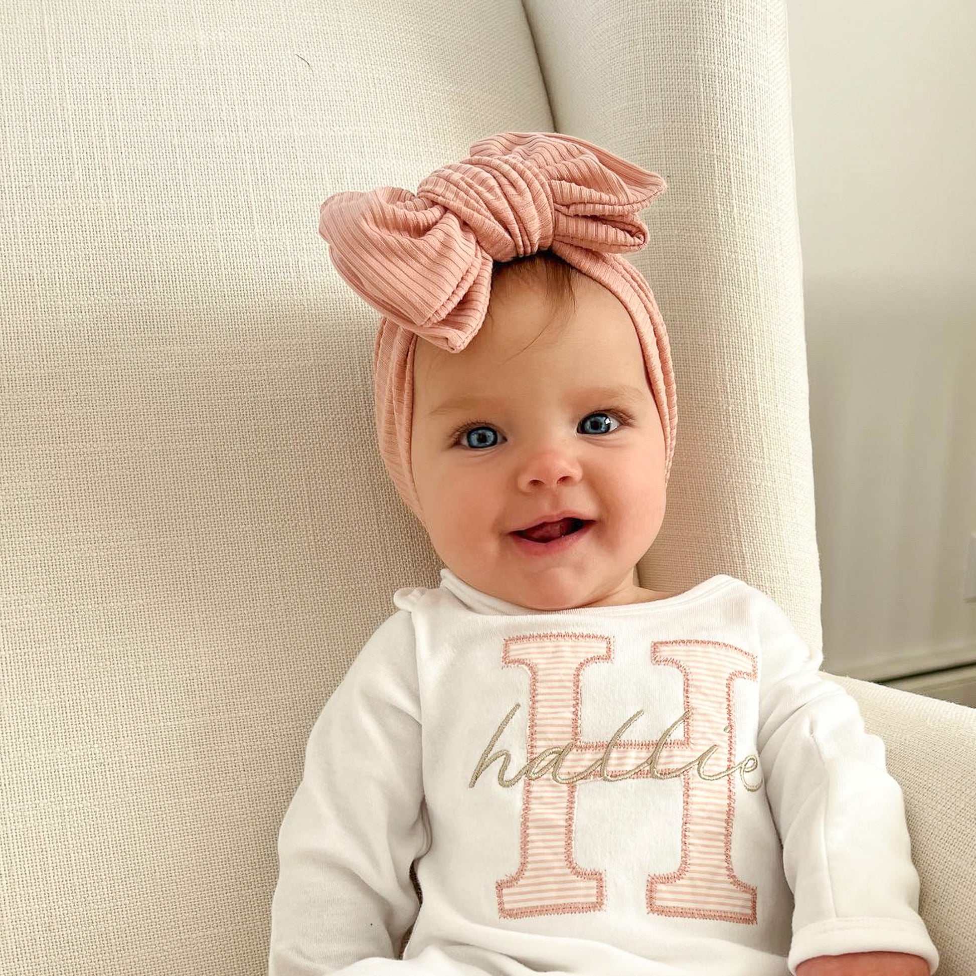Baby wearing a dusty pink headband and a white outfit with 'Hattie' printed on it, sitting on a beige couch.