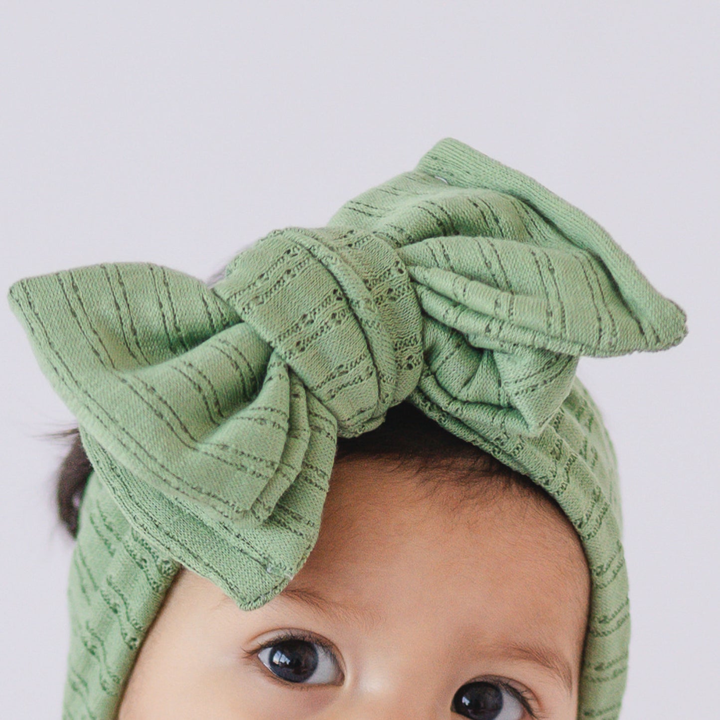 Close up view of baby wearing a green textured oversized bow headband on a light gray background