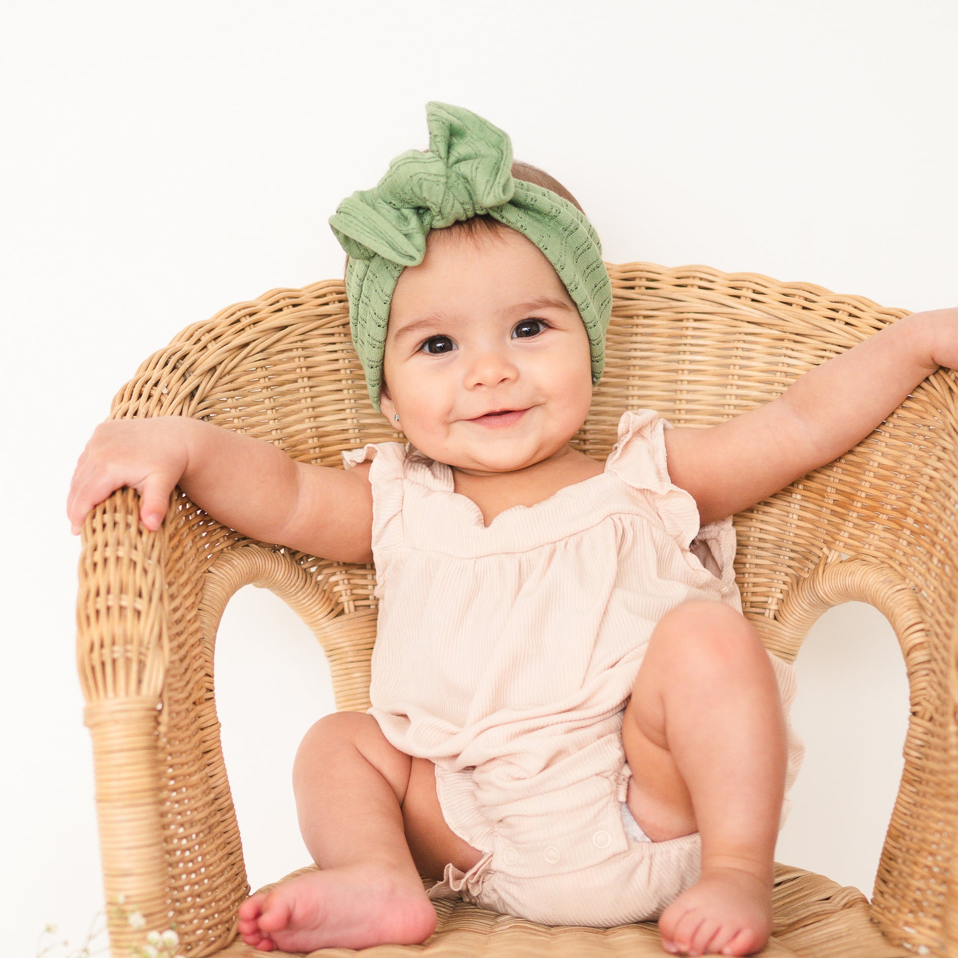 Baby sitting on a wicker chair wearing a green headband and beige outfit.