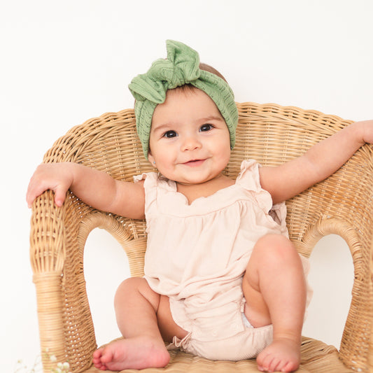 Baby sitting on a wicker chair wearing a green headband and beige outfit.