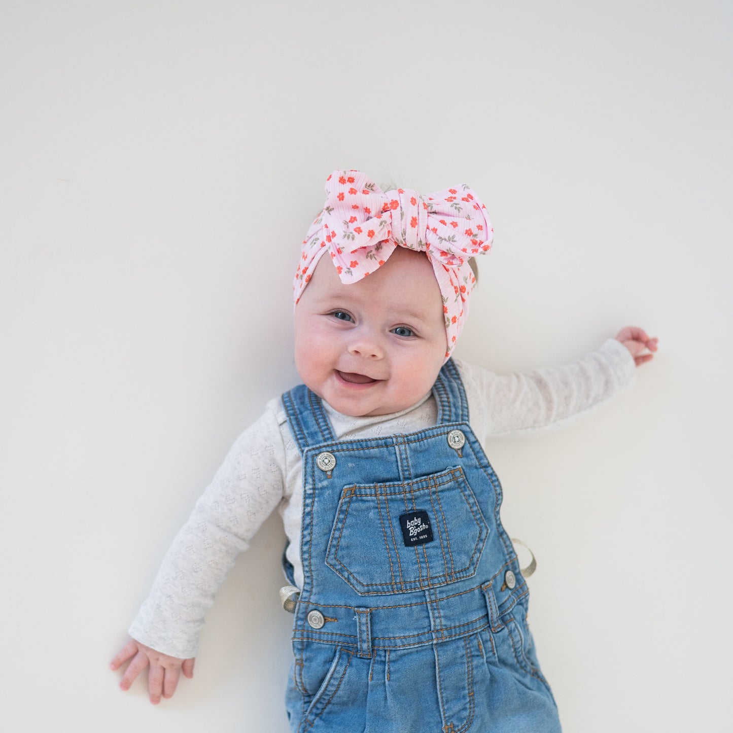 Baby wearing denim overalls and a floral headband on a plain background