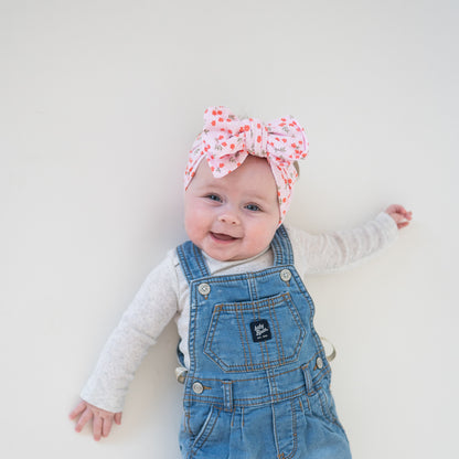 Baby wearing denim overalls and a floral headband on a plain background