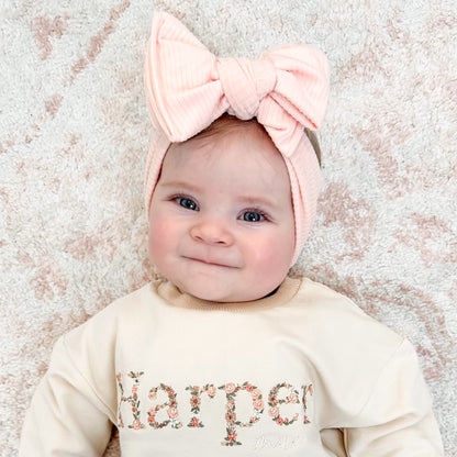 Baby wearing a pink headband with a bow and a sweater with 'Harper' printed on it, against a textured beige background.