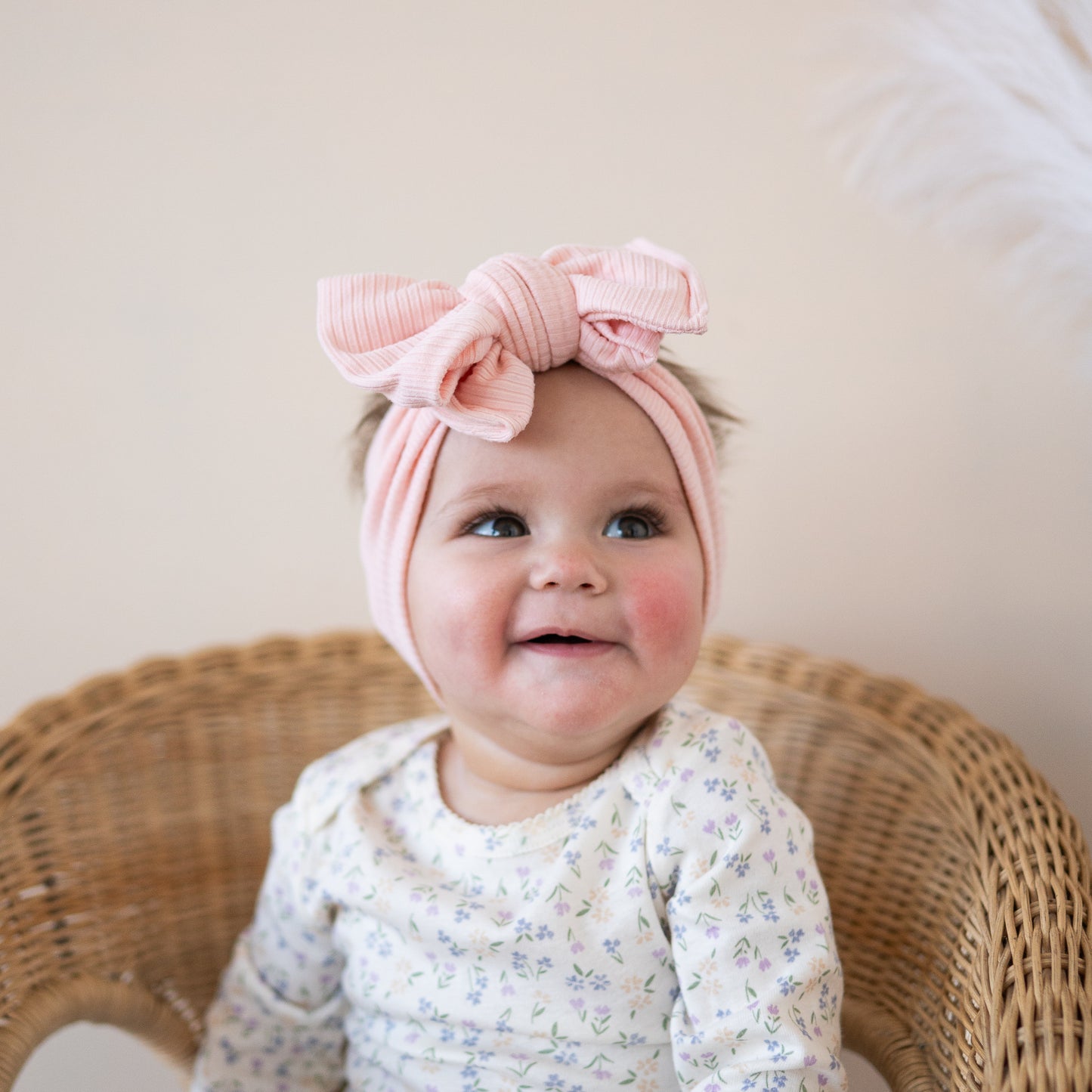 Baby wearing a pink headband with a bow, sitting on a wicker chair.
