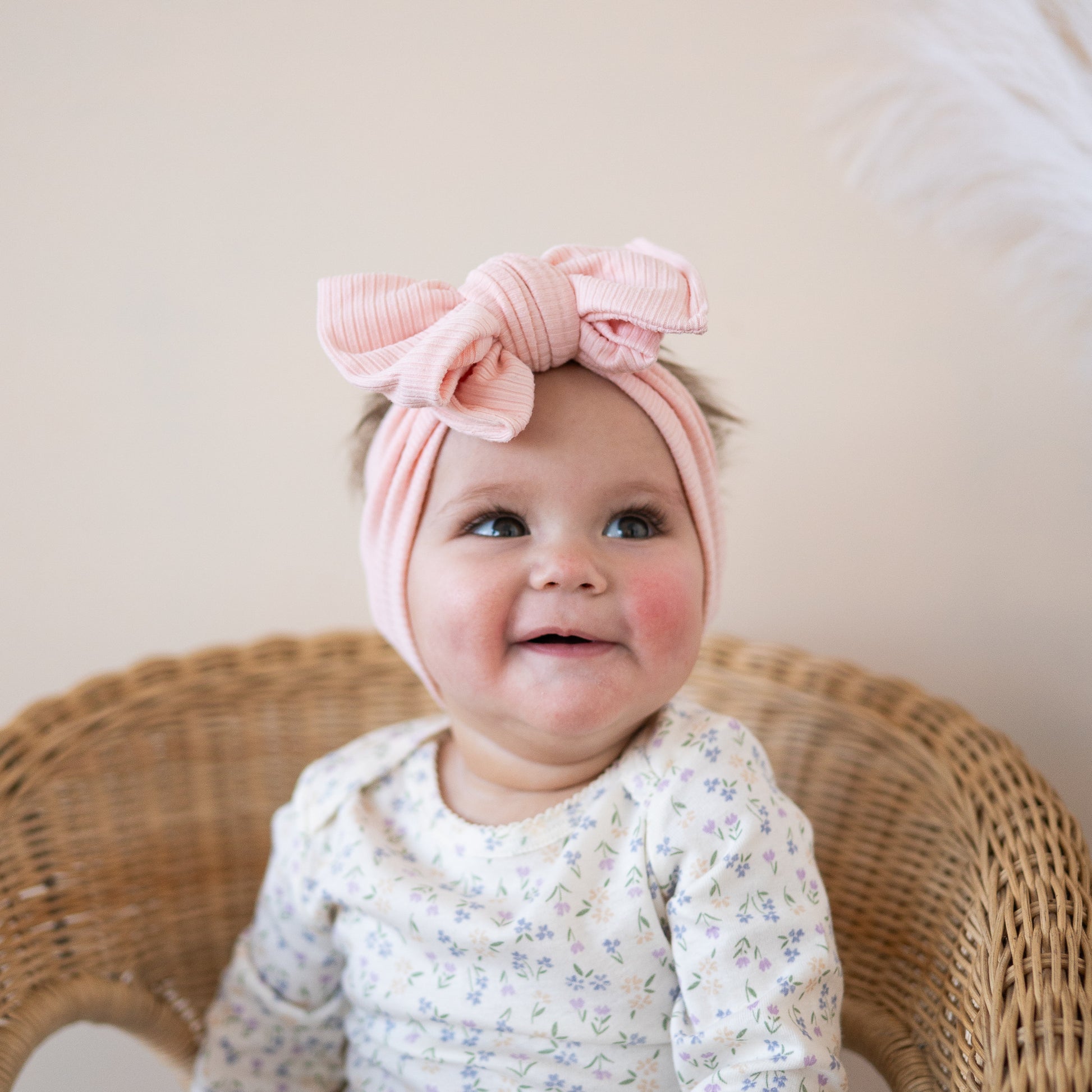 Baby wearing a pink headband with a bow, sitting on a wicker chair.