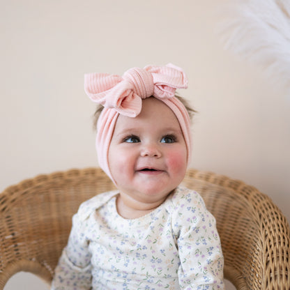 Baby wearing a pink headband with a bow, sitting on a wicker chair.