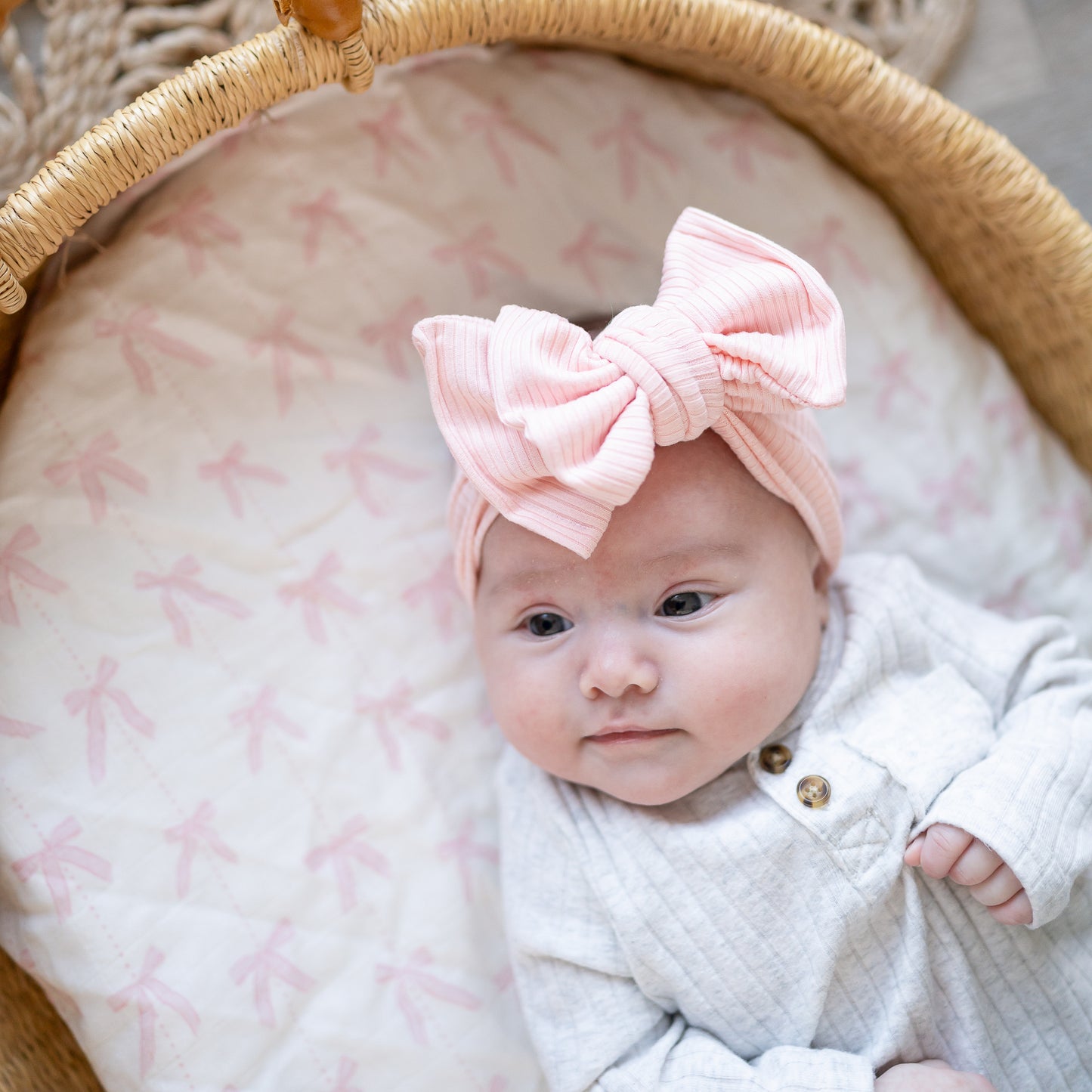Newborn baby wearing a pink oversized bow headband in Moses basket with pink and white sheets.