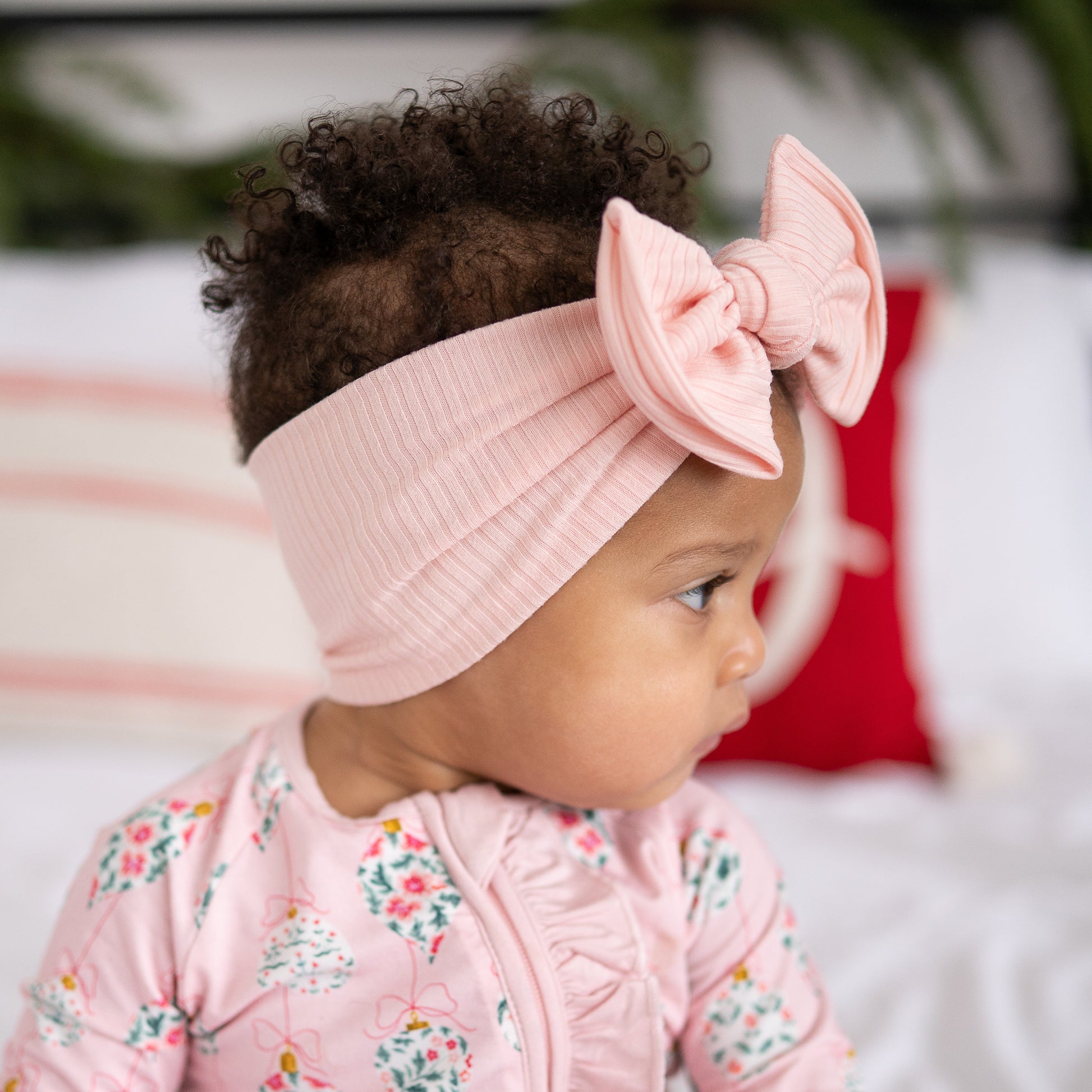 Side view of a baby wearing a pink ribbed headband with a top knot bow, sitting on a bed.