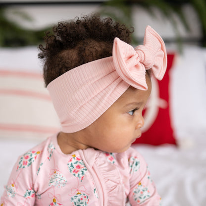 Side view of a baby wearing a pink ribbed headband with a top knot bow, sitting on a bed.