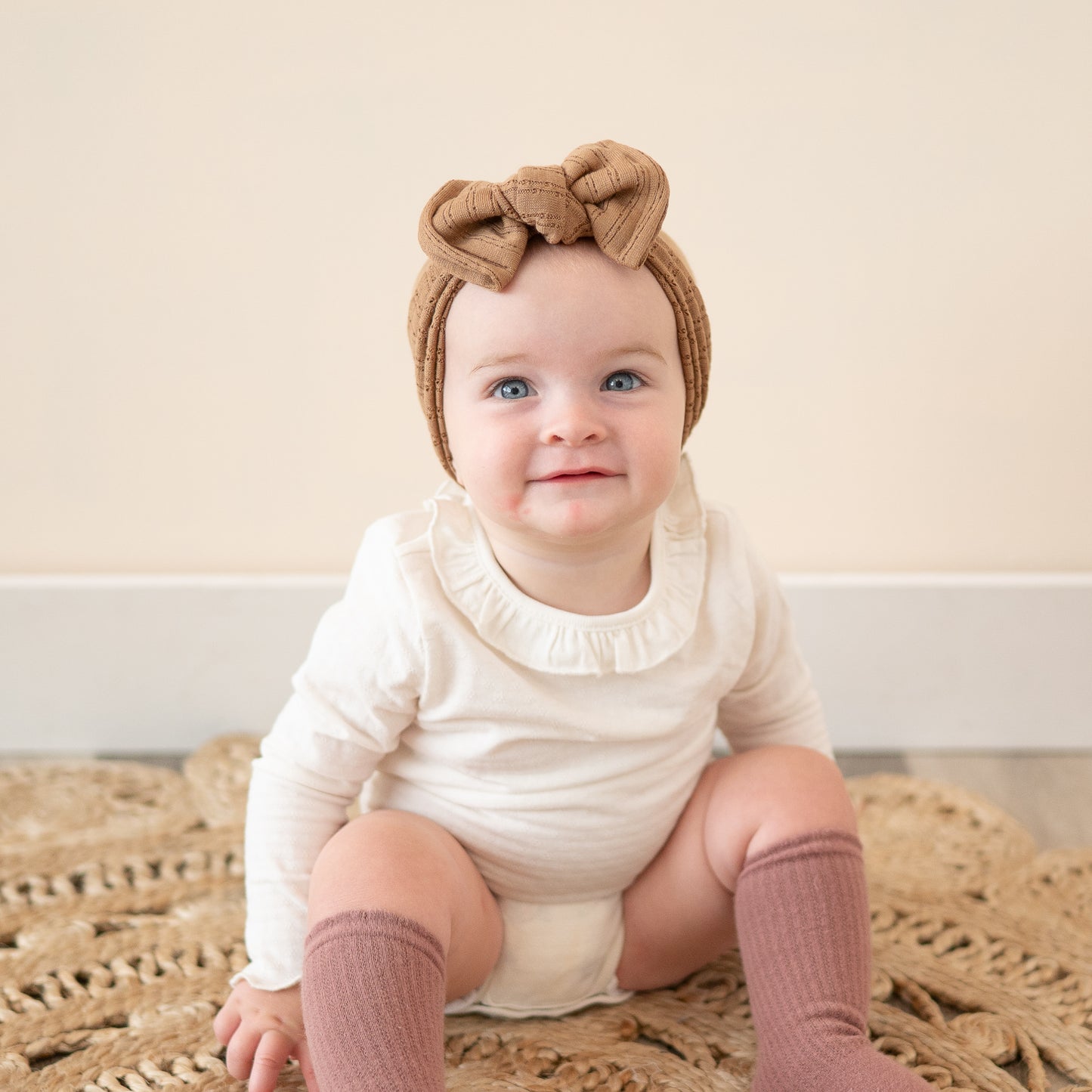 Baby smiling and wearing a mocha brown pointelle headband with a top knot, white onesie, and pink socks sitting on a textured rug.