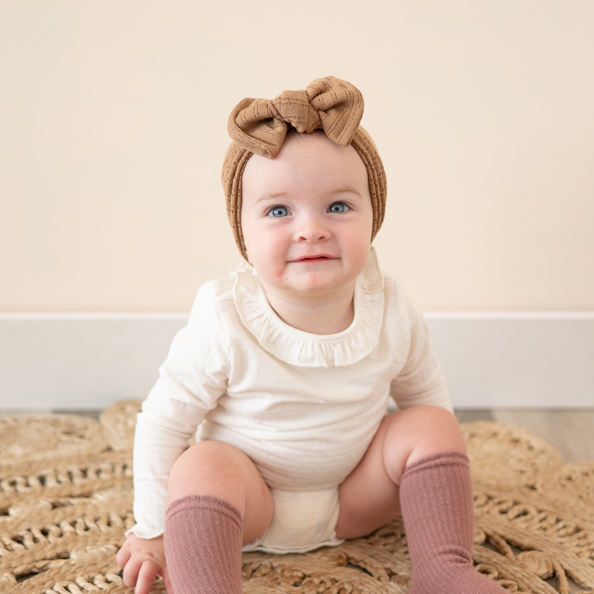 Baby smiling and wearing a mocha brown pointelle headband with a top knot, white onesie, and pink socks sitting on a textured rug.