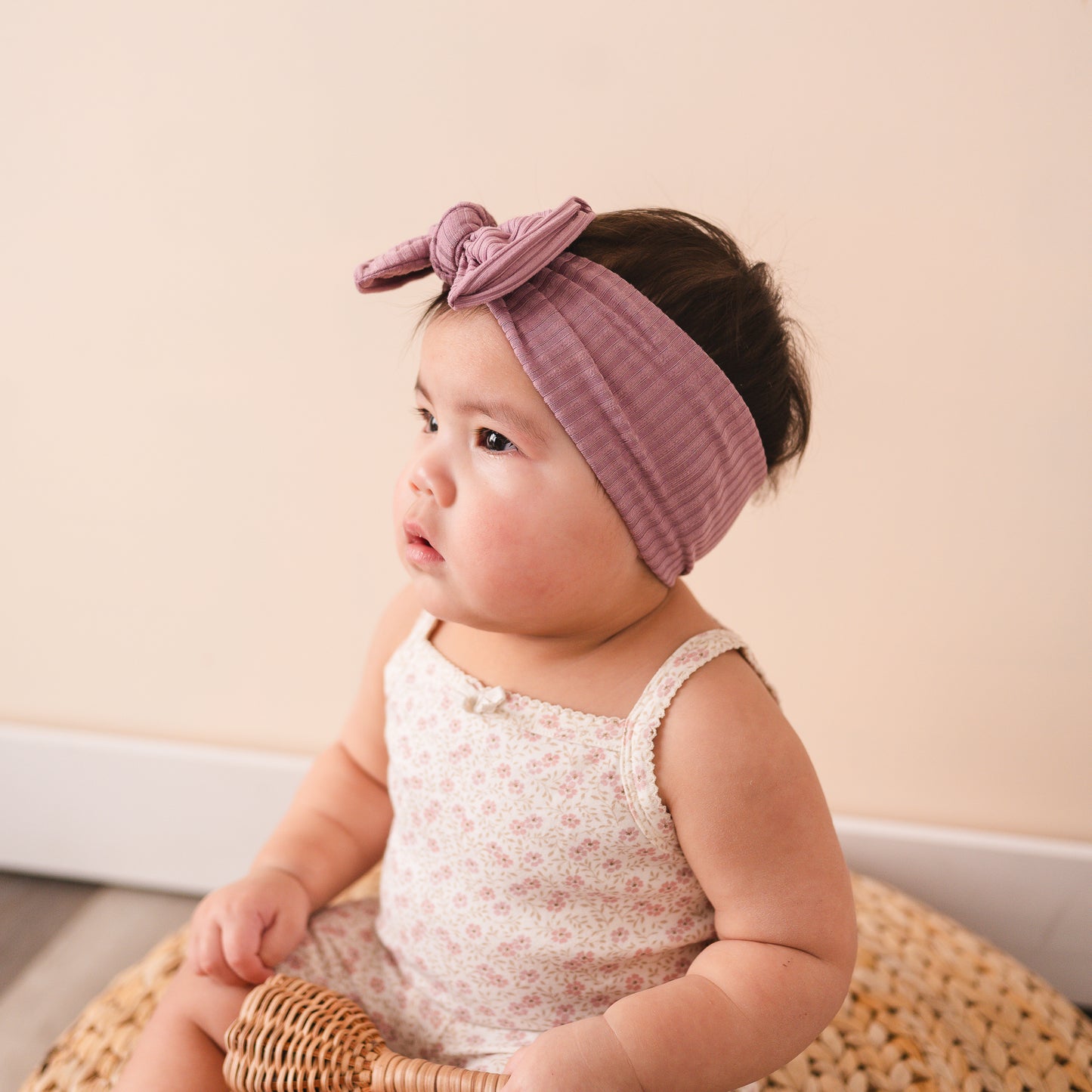 Baby wearing a purple headband sitting on a woven pouf with a plain background