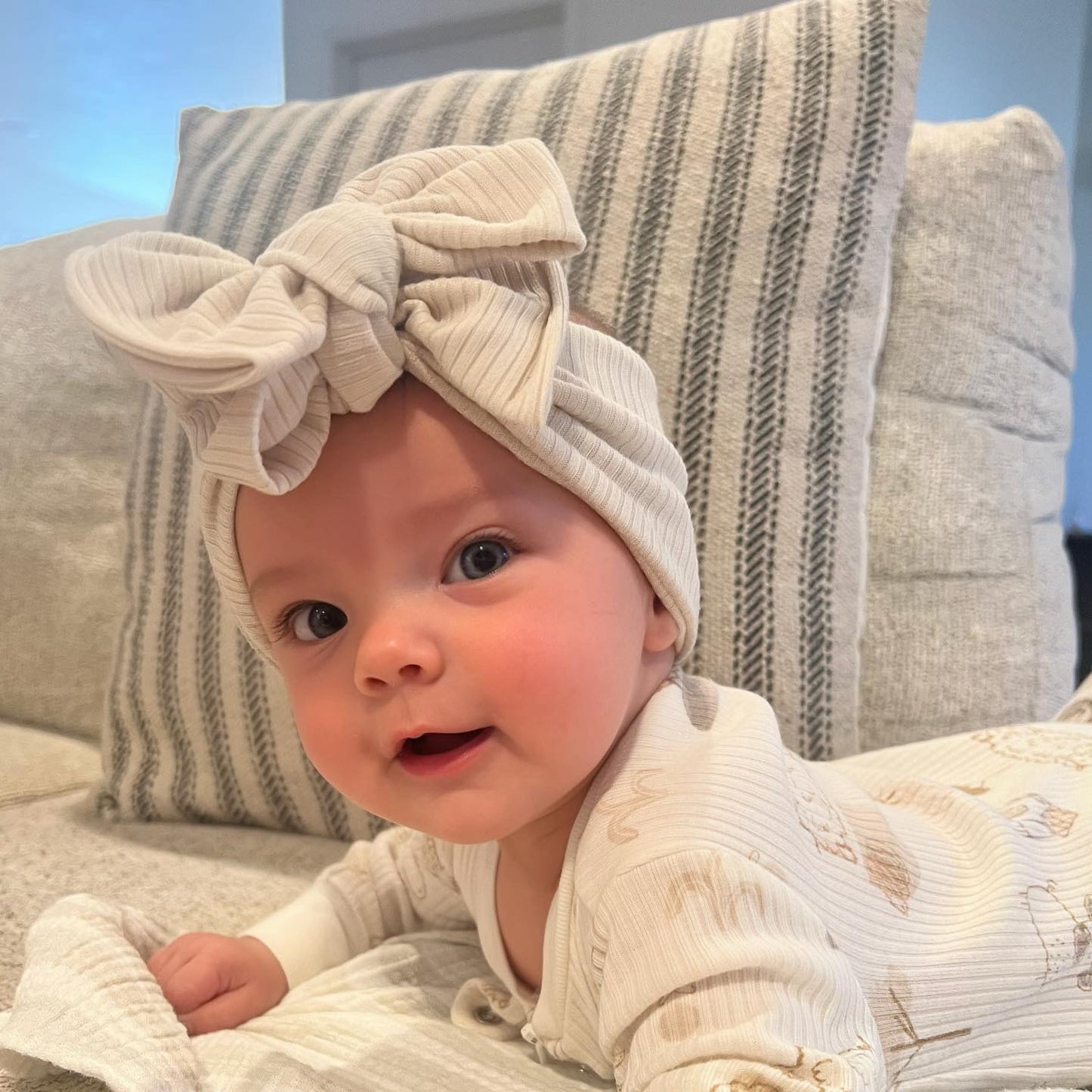 Baby wearing a beige ribbed headband with an oversized bow, sitting on a couch.