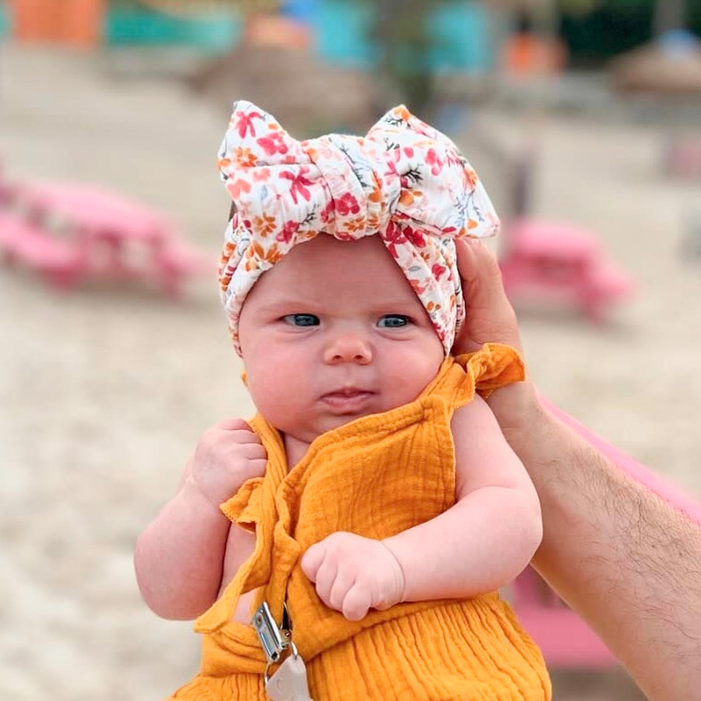 Baby in a yellow outfit with a floral headband held by an adult at a playground.