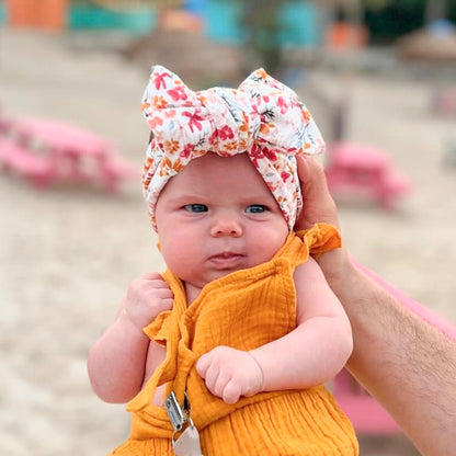 Baby in a yellow outfit with a floral headband held by an adult at a playground.
