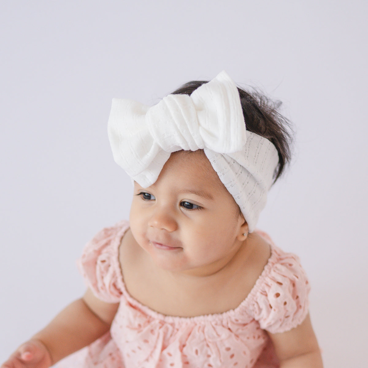 Baby wearing a pink dress and white pointelle oversized bow headband on a light background