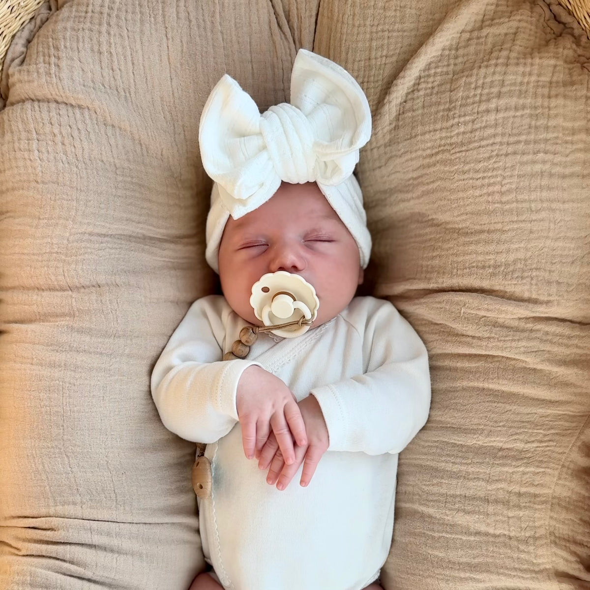 Newborn baby wearing a white outfit and headband, lying on a textured beige surface.