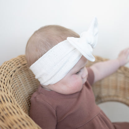 Baby wearing a white headband with a bow, sitting on a wicker chair.
