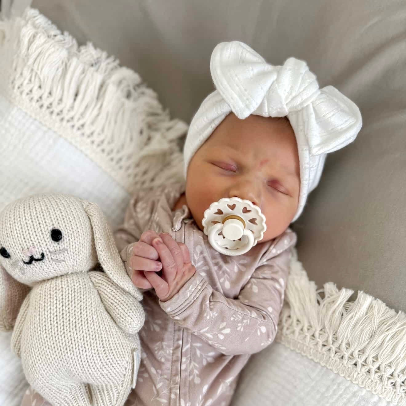Newborn baby wearing a white bow headband and holding a cuddle + kind doll, with a neutral background.