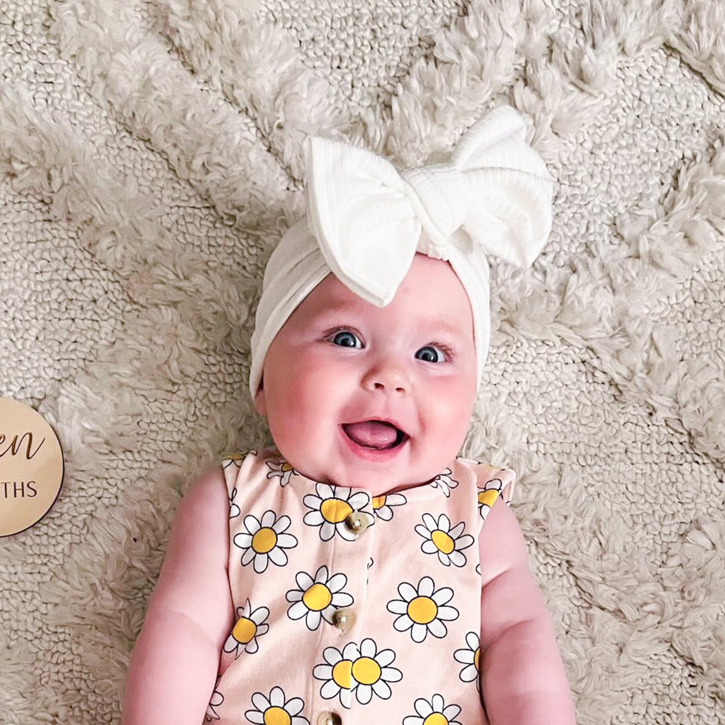 Baby wearing a white headband with a bow, lying on a soft beige surface.