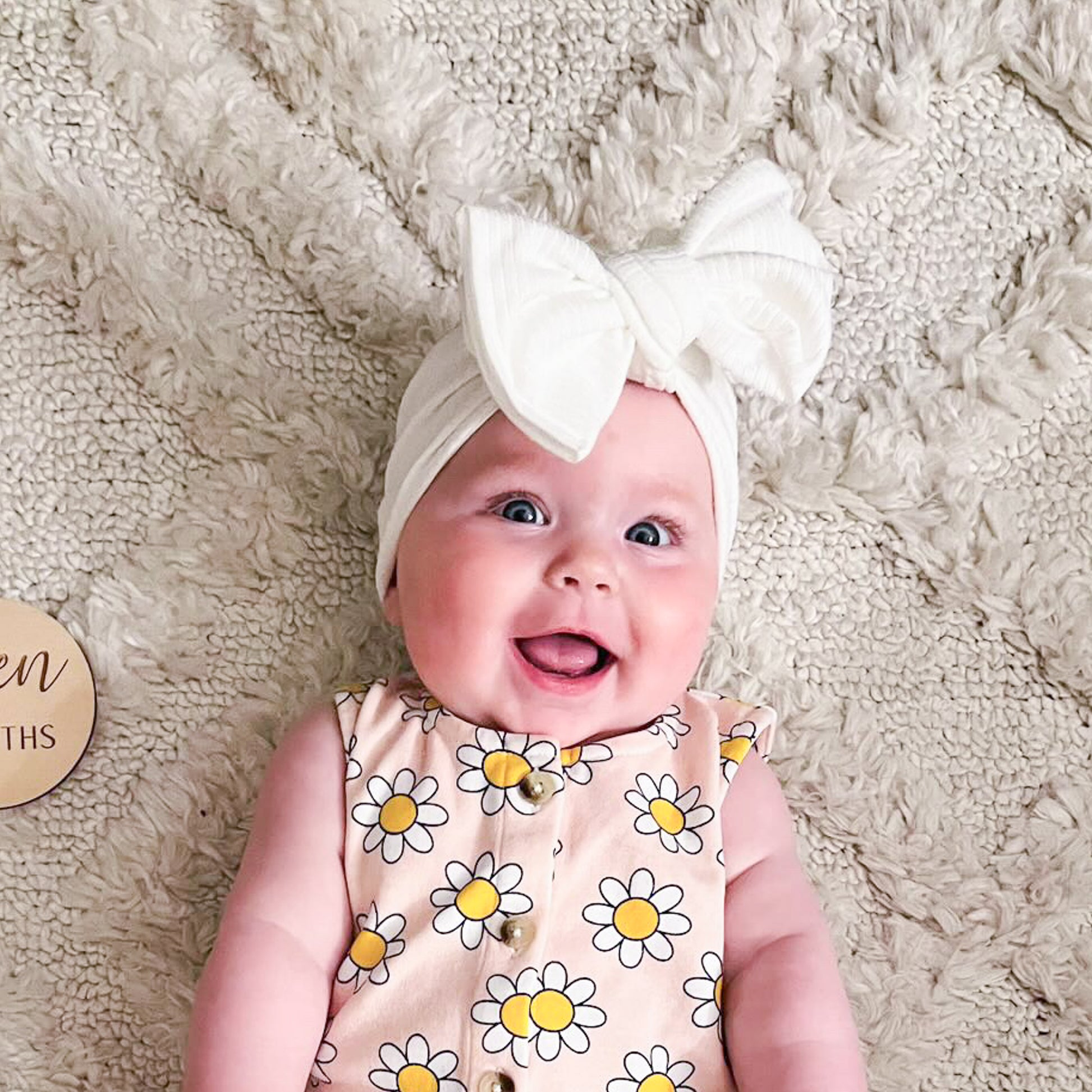 Baby wearing a white headband with a bow, lying on a soft beige surface.