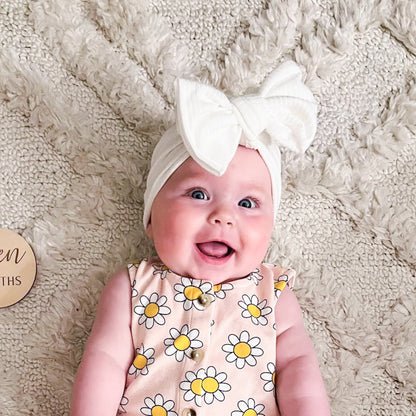 Baby wearing a white headband with a bow, lying on a soft beige surface.