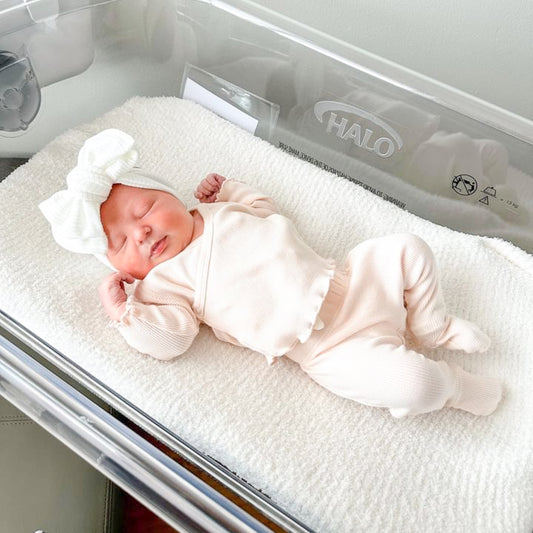 Newborn baby girl in a hospital cot wearing a white ribbed oversized bow.