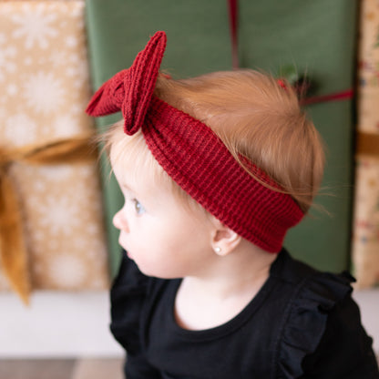 Child wearing a red knitted headband with a bow, standing in front of gift-wrapped presents.
