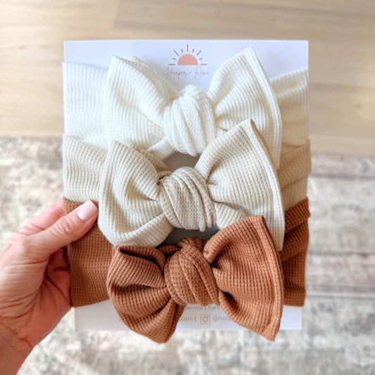 Set of three neutral waffle knit baby oversized bow headbands, held by a hand.