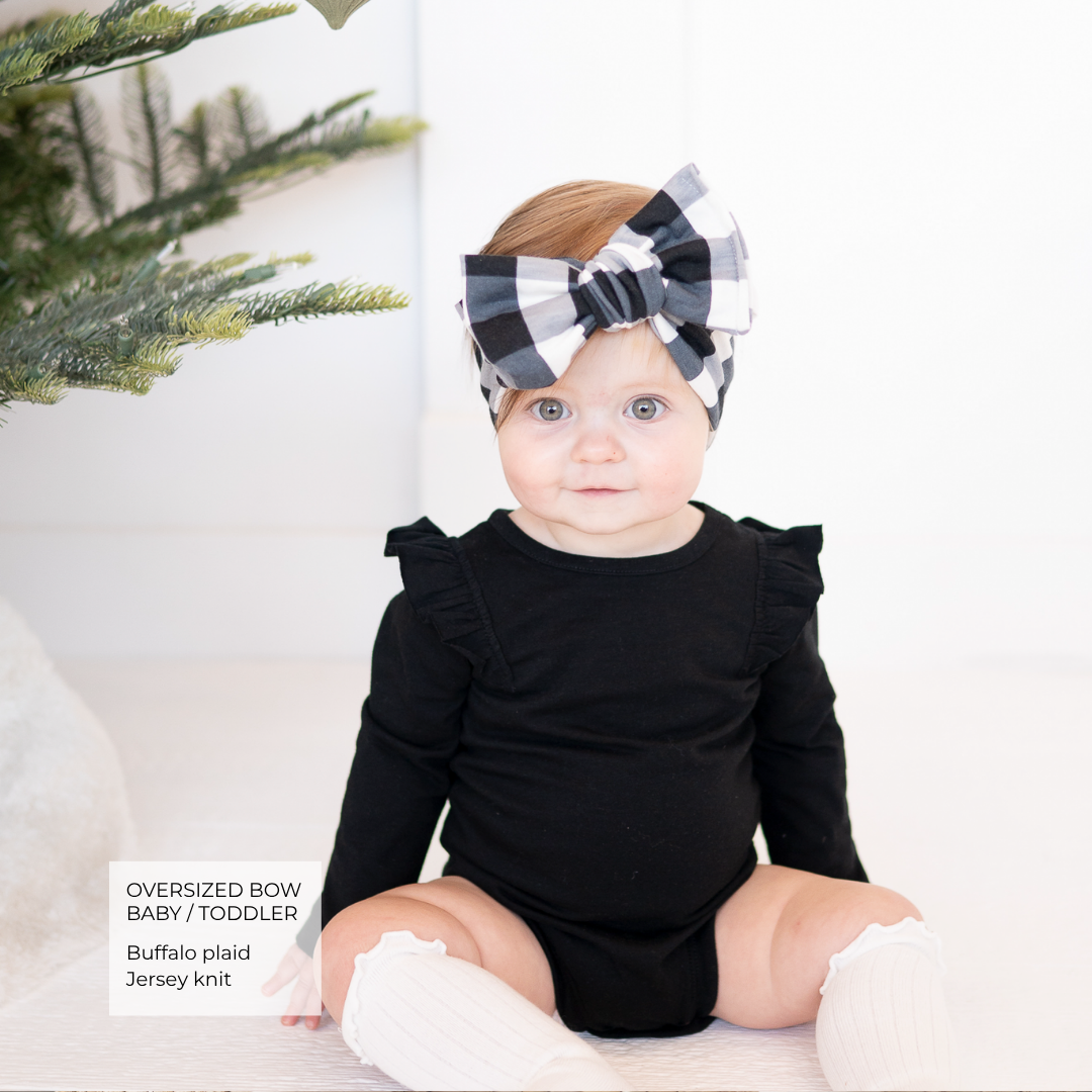 A baby wearing a black long-sleeve bodysuit with ruffled shoulders, white knit socks, and a large buffalo plaid bow headband, sitting in front of a white background with a Christmas tree branch visible on the left.