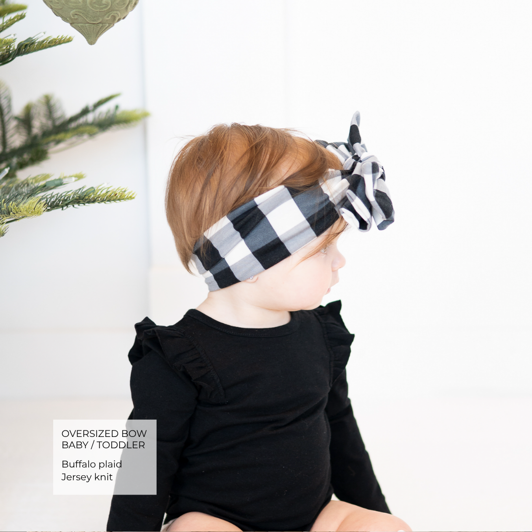 A toddler wearing an oversized buffalo plaid bow headband and a black long-sleeve top, shown in profile against a bright white background with a small green plant visible at the left edge.