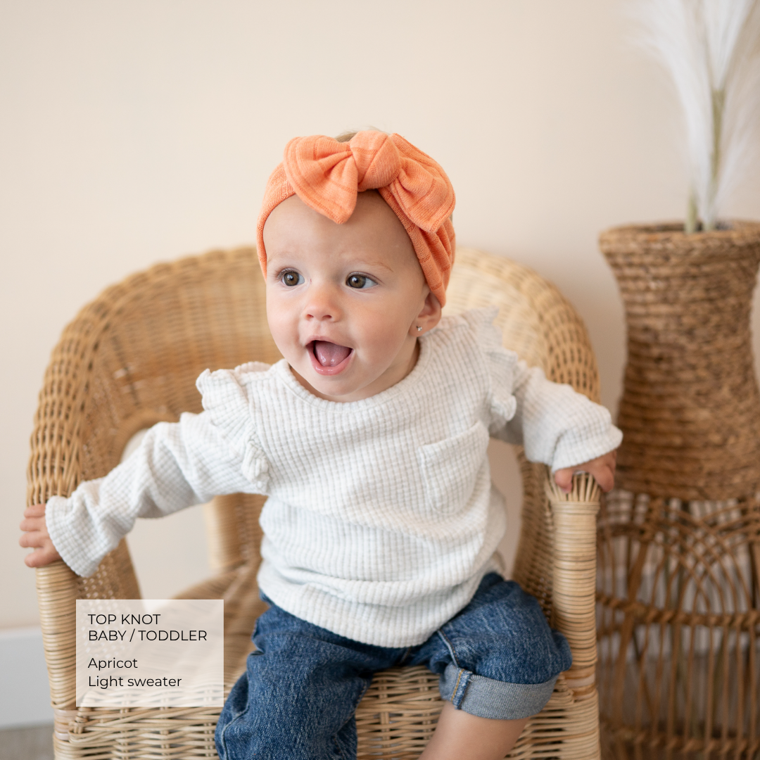 A baby wearing a peach apricot knit top knot headband and a white waffle-knit long-sleeve sweater with ruffle trim, sitting in a woven rattan chair. The image includes a text overlay on the bottom left identifying the product as a 'Top Knot Baby / Toddler Apricot Light sweater'.