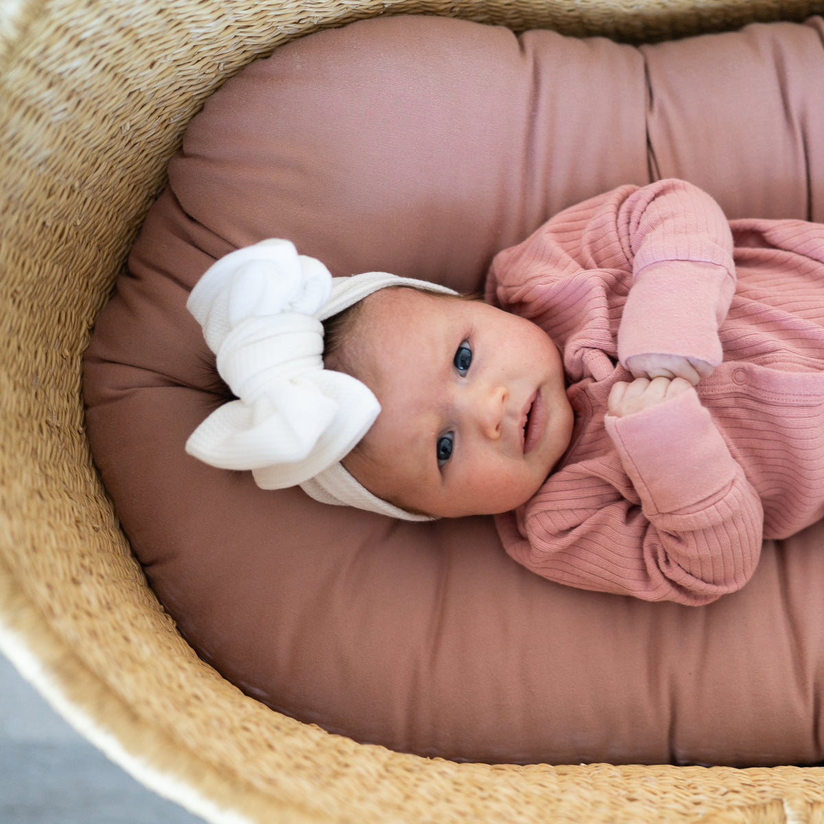 Newborn baby in a pink outfit and white headband lying on a brown cushion in a woven basket.
