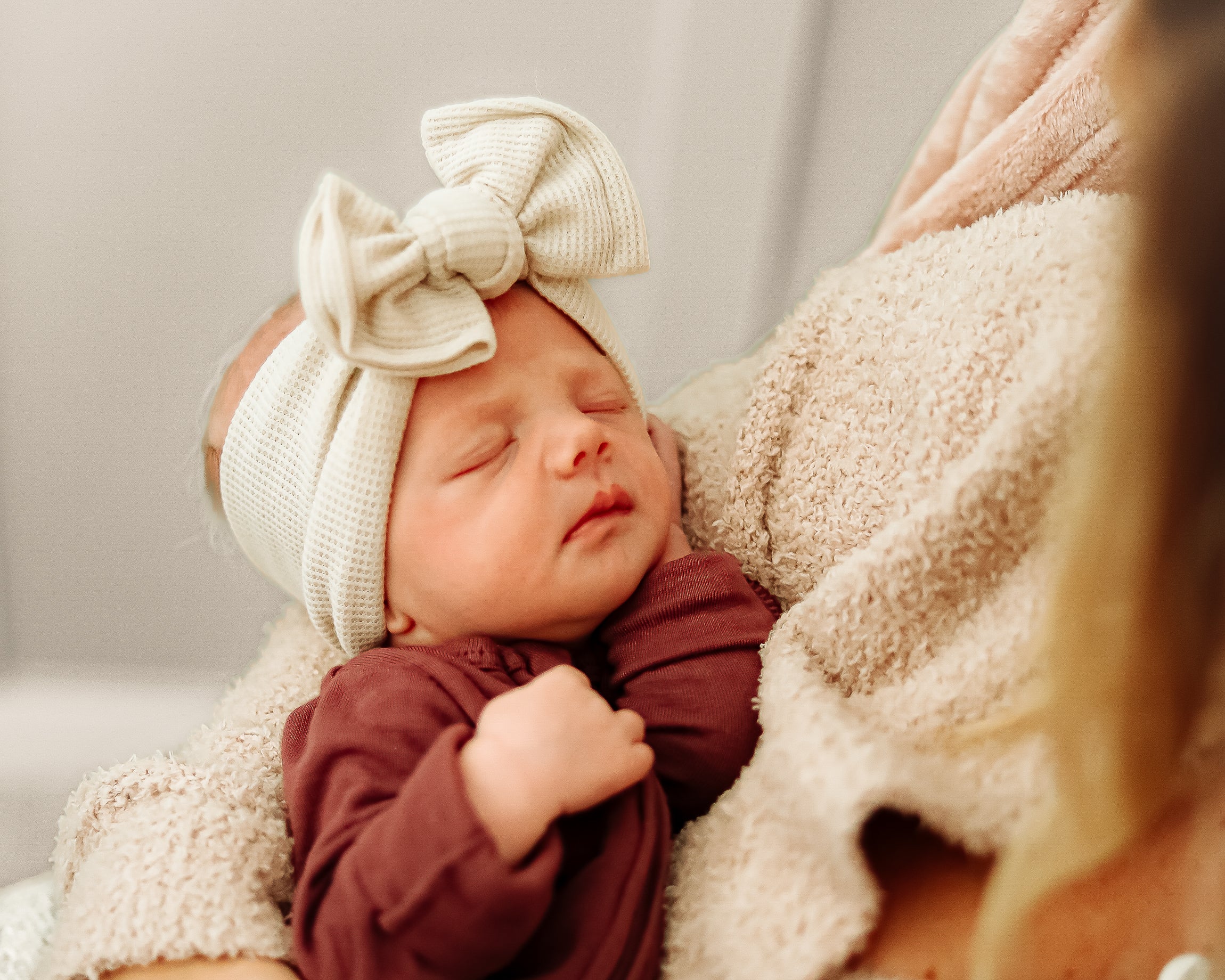 Newborn baby wearing a cream headband with a bow, wrapped in a brown outfit and beige blanket.
